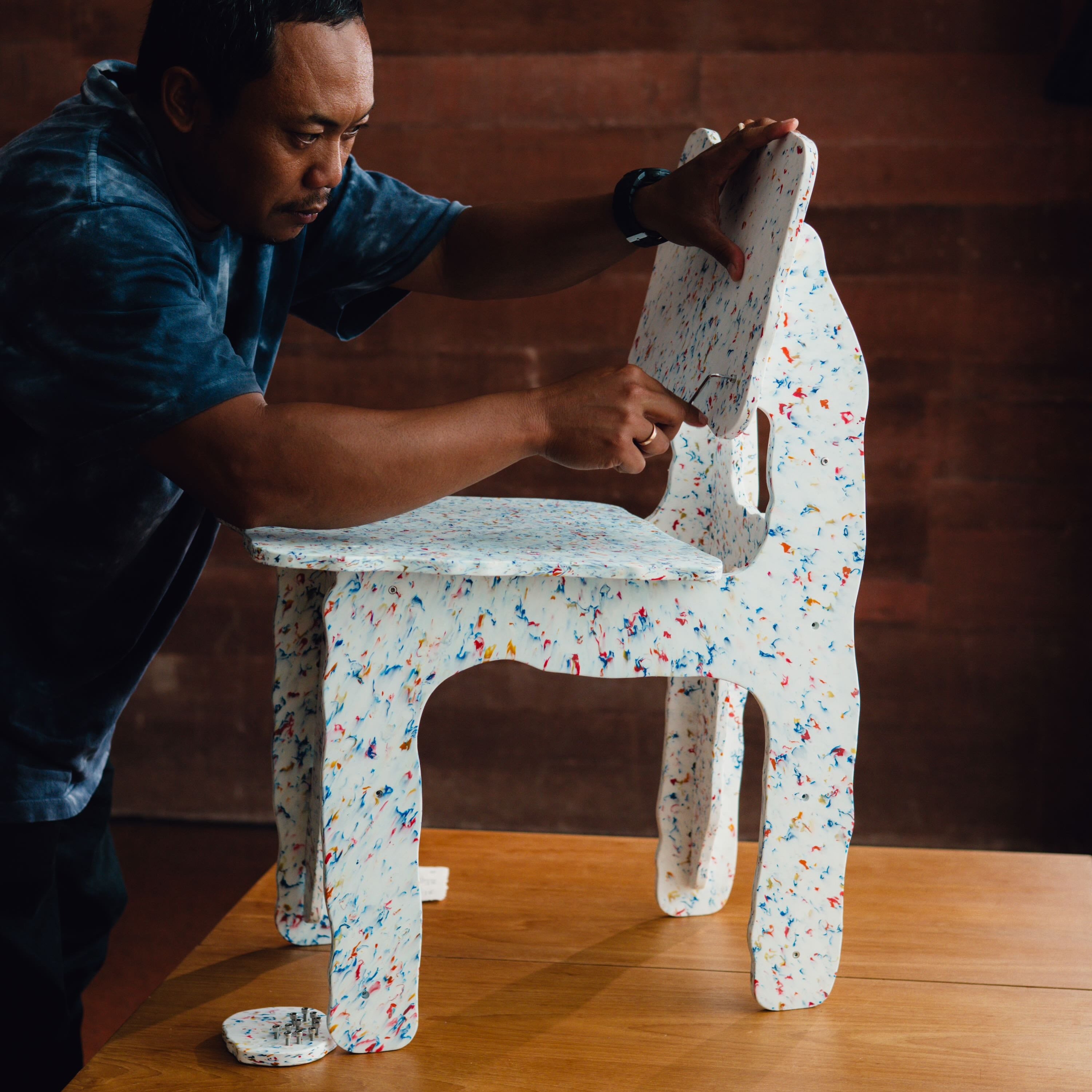 Person adjusting a small, colorful chair on a wooden surface with a wooden wall background