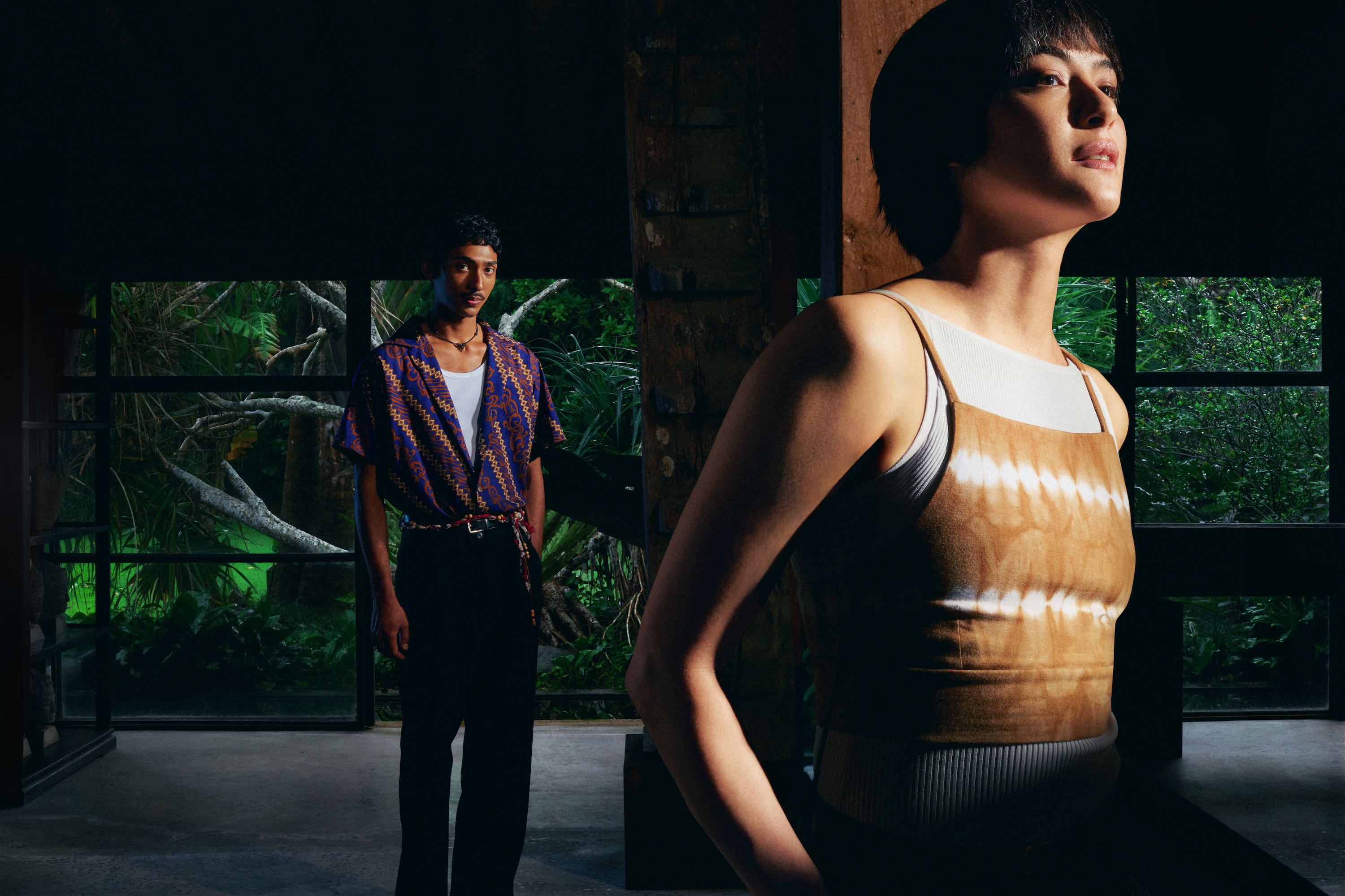 Two people standing in a room with greenery visible through windows.