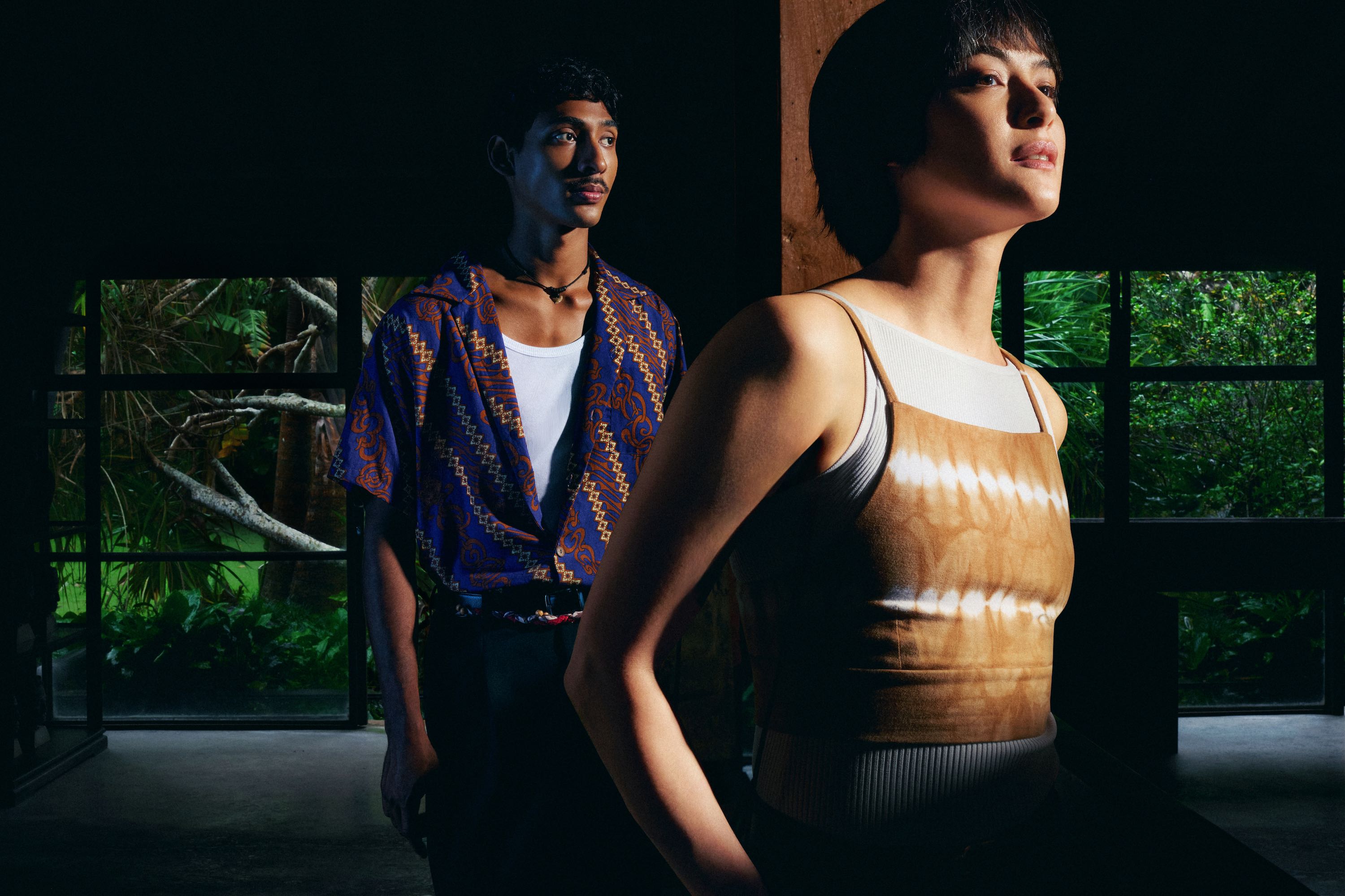 Two people standing in a room with large windows showing greenery outside.